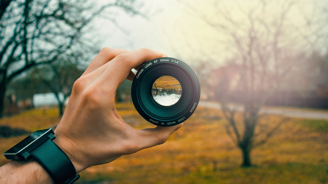 A hand holding a camera lens outdoors, with a blurred natural background in autumn colors.