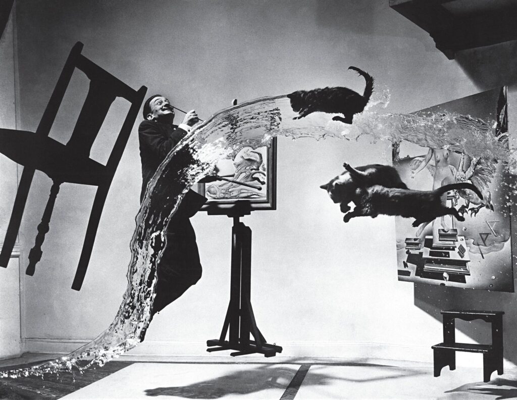 Surreal black-and-white photograph featuring cats, water, and a man posing in mid-air with a chair and painting in the background.