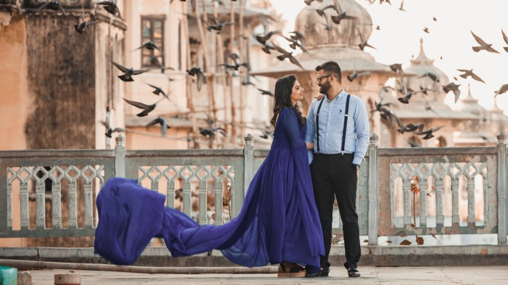 Couple posing in a romantic setting with birds flying in the background and a flowing blue dress on a historic balcony.