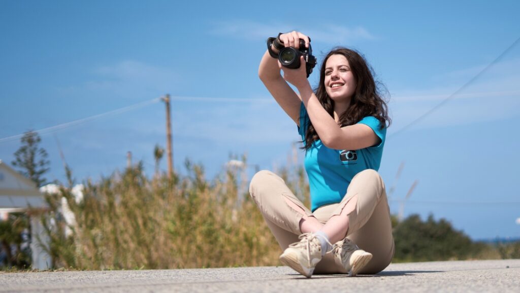 Smiling woman sitting outdoors holding a camera, enjoying a sunny day with a natural background.