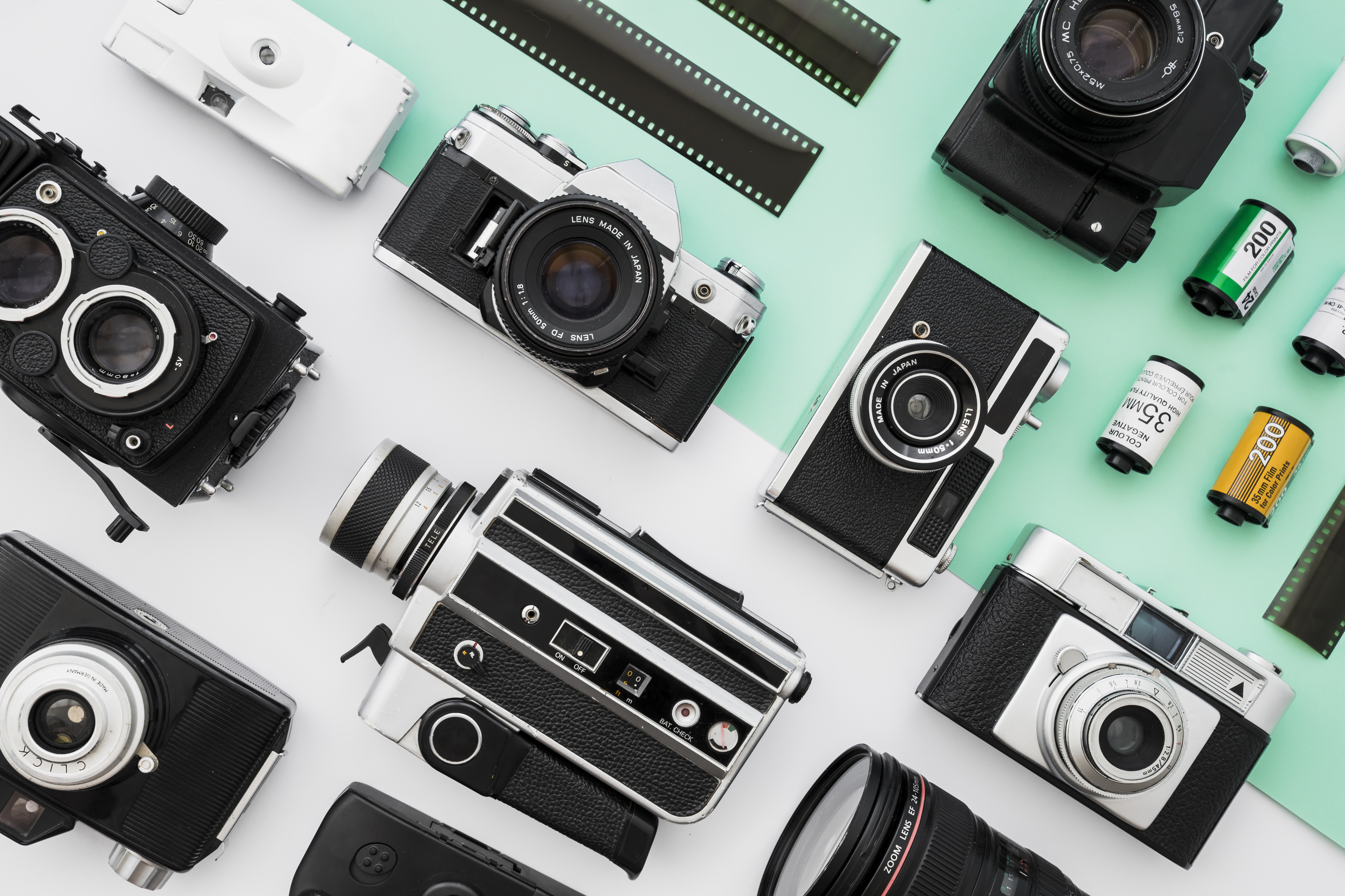 Collection of vintage photo and video cameras on a teal background, with film rolls nearby.