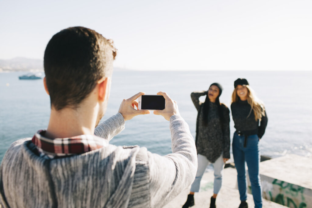 Man taking a photo of two smiling women posing by the sea on a sunny day.